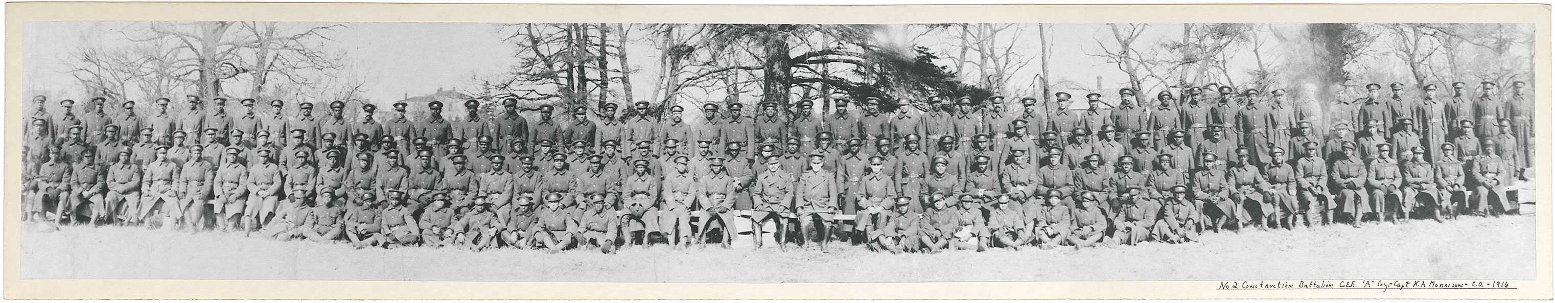 Black and white photograph. A large group of majority black men sit for a group photo. Most are wearing long military coats. Trees and rooftops are visible in the background.